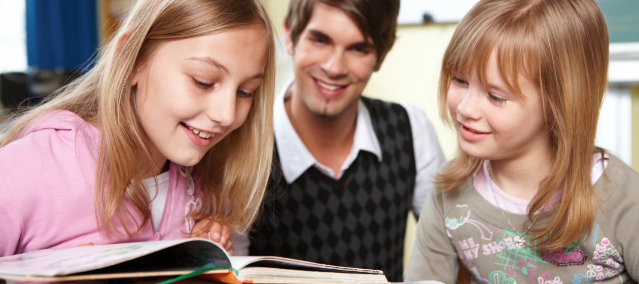 Zwei Schülerinnen lesen etwas in einem Buch, der Lehrer sitzt hinter ihnen und lächelt