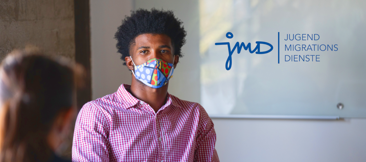 Ein junger Mann mit buntem Nase-Mund-Schutz spricht mit einer Kollegin vor dem Logo der Jugendmigrationsdienst JMD.