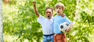 Zwei Jungen unterschiedlicher Herkunft stehen dicht nebeneinander und mit einem Fussball im Park und strecken die Faust in die Luft und rufen etwas.