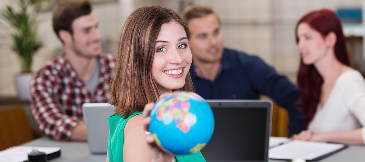 Junge Frau sitzt lächelnd mit einer Gruppe an einem Tisch und hält einen Globus in die Kamera. 