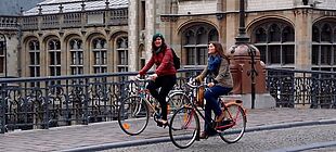 Zwei junge Frauen fahren mit Rädern über eine Steinbrücke in der Genter Altstadt.