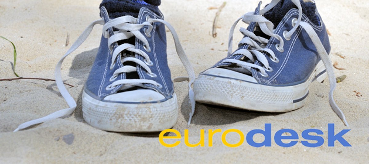Ein paar blaue Schuhe mit weißer Sohle und weißen Schnürsenkeln ("Chucks") stehen auf Sand. In der Ecke rechts unten befindet sich das Logo von Eurodesk. 