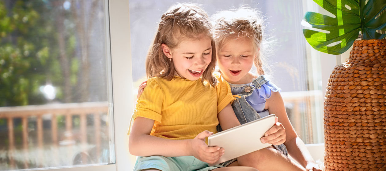 Zwei Mädchen im Grundschul- und Kindergartenalter sitzen zu Hause vor einem Tablet und lachen.