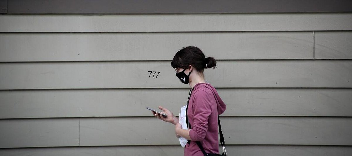 Frau in einem Hoodie und Mundschutz hält Papiere in ihrer Hand und schaut auf ihr Smartphone
