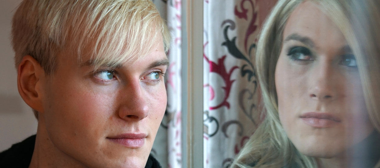 Ein junger Mann schaut in eine Fensterscheibe und sein Spiegelbild ist weiblich. 