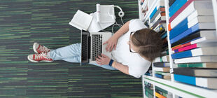 Ein Student sitzt in einer Bibliothek mit seinem Laptop und Unterlagen auf dem Boden und arbeitet.