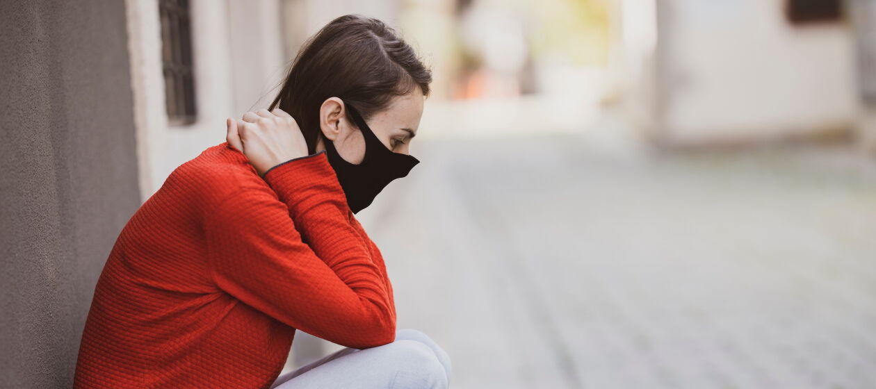 Eine junge Frau trägt eine Atemschutzmaske und lehnt in der Hocke verzweifelt an einer Häuserwand