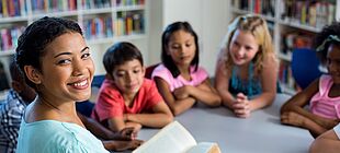Mehrere Kinder sitzen in einer Bücherei um einen Tisch, eine erwachsene Person liest aus einem Buch vor