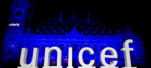 Gebäude wird blau angestrahlt, davor steht eine Skulptur mit den Buschtaben UNICEF