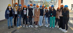 Eine Gruppe Jugendlicher steht mit Atemschutzmasken und Sicherheitshelmen auf dem herbstlich kalten Werksgelände einer Chemiefabrik.