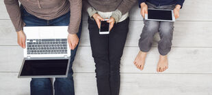 Eine Familie sitzt nebeneinander auf einem Holzboden und alle schauen auf Laptop,Smartphone oder Tablet. 