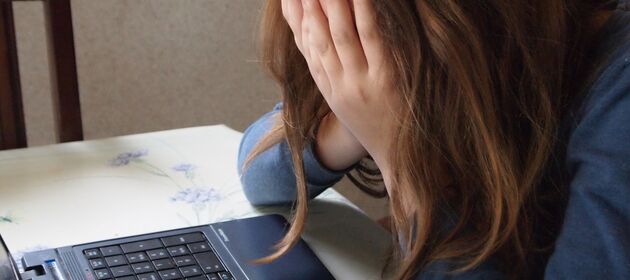 Eine Jugendliche sitzt vor dem Laptop und hält mit geneigtem Kopf verzweifelt die Hände vors Gesicht