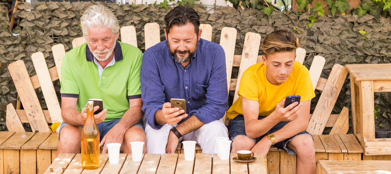 Großvater, Sohn und Enkel sitzen nebeneinander auf einer Bank im Freien und schauen alle drei auf ihr jeweils eigenes Smartphone