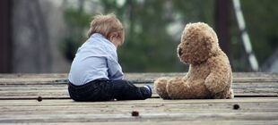 Ein Kleinkind sitzt mit einem Teddybär ihm gegenüner alleine und mit gesenktem Kopf auf einer Holzbrücke