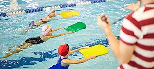 Drei Kinder schwimmen mit Schwimmbrettern in einem Schwimmbecken, eine erwachsene Person steht am Beckenrand