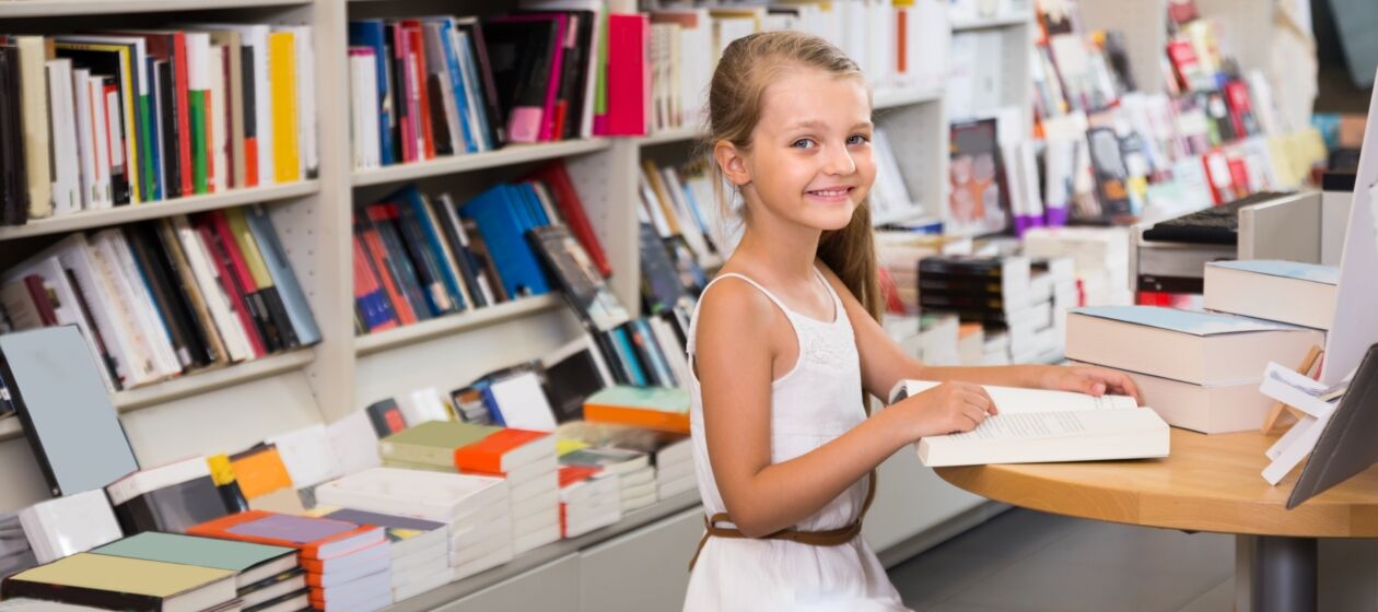 lächelndes Mädchen mit aufgeschlagenem Buch an einem Tisch in Buchhandlung