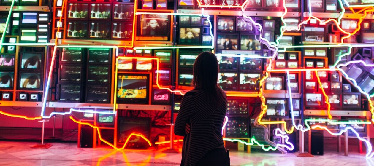 Eine junge Frau betrachtet in einem Museum eine Installation mit vielen bunten Lichtern und technischen Gegenständen