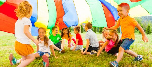 Eine Gruppe freudiger Kinder versteckt sich auf einer Wiese unter einem regenbogenfarbenen Tuch