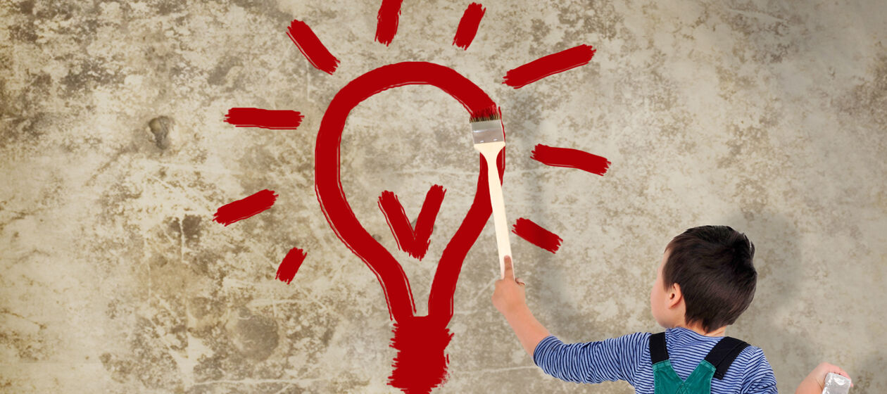 Ein Junge mit latzhise malt eine Glühbirne in roter Farbe an eine Wand