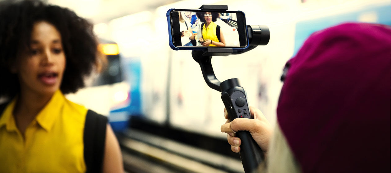 Zwei junge Frauen stehen am Gleis in einem Bahnhof und filmen mit dem Smartphone.
