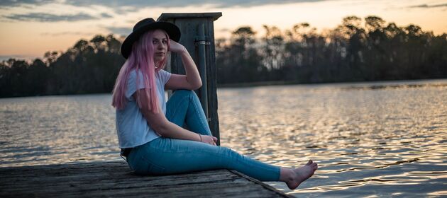 Jugendliche mit rosa Haaren und Hut auf einem Steg am See bei rosarotem Abendhimmel