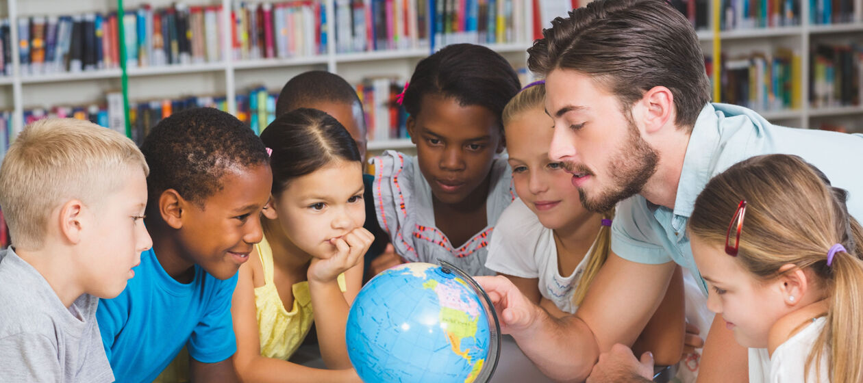 Schüler/-innen schauen gemeinsam mit ihrem Lehrer in einer Bibliothek auf einen Globus. 