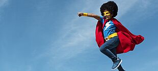 Ein als Superheld verkleideter dunkelhäutiger Junge springt kraftvoll in die Luft