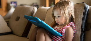 Ein kleines Mädchen sitzt zu Hause auf der Couch und schaut gebannt auf ein Tablet.