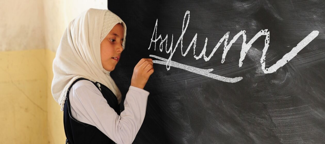 Ein Mädchen mit Kopftuch schreibt auf eine Tafel das englische Wort Asylum