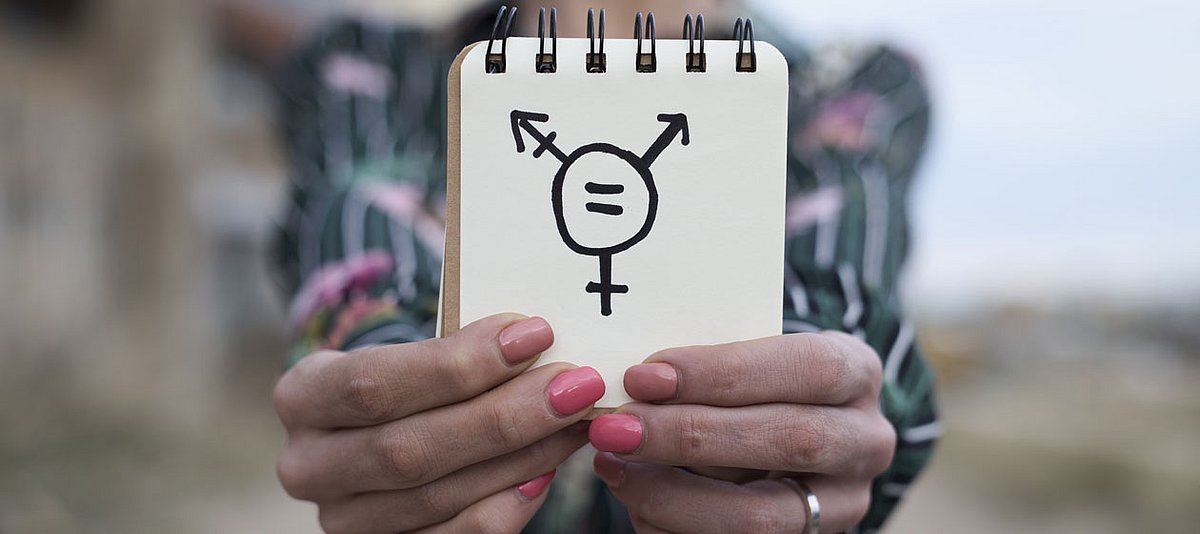 Eine Frau hält einen Notizblock in den Händen, auf dem das Transgender Symbol gemalt ist.