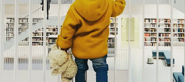 Ein Kind mit einem Teddy in der Hand schaut auf viele Regale voller Bücher