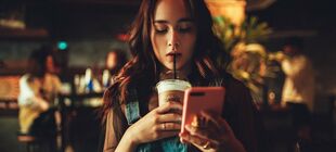 In einem Café schlürft eine junge Frau am Strohhalm ihre Kaffeegetränks. Sie schaut auf das Telefon in ihrer Hand.