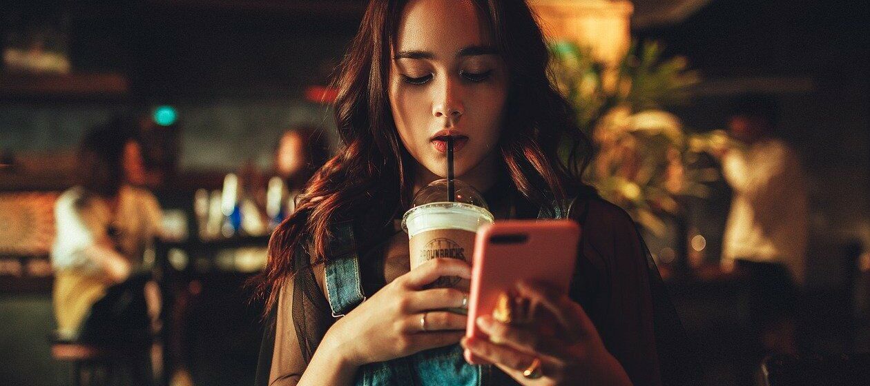 In einem Café schlürft eine junge Frau am Strohhalm ihre Kaffeegetränks. Sie schaut auf das Telefon in ihrer Hand.
