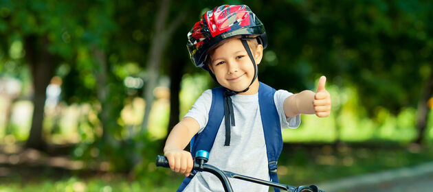 Eine Junge mit Fahrradhelm sitzt auf einem Fahrrad, streckt den Daumen hoch und lächelt.   