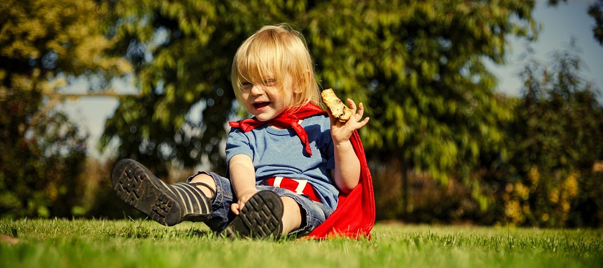 Ein kleines Kind sitz mit Superheldenumhang auf eier Wiese, lacht und hält etwas zu essen in der Hand