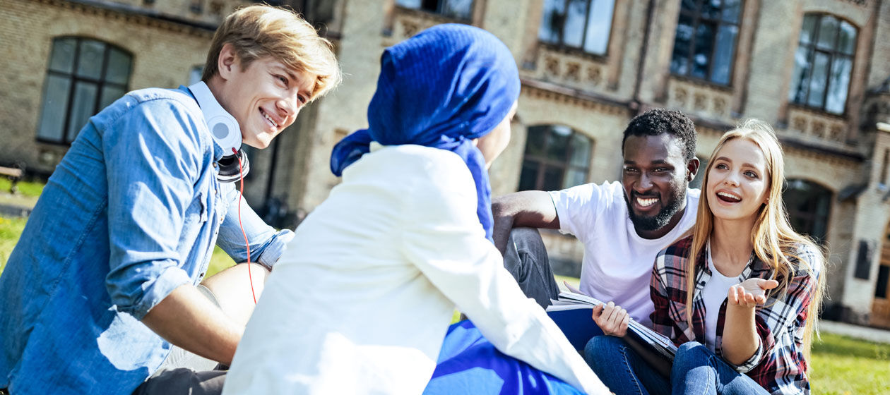 Studenten unterschiedlicher Herkunft sitzen lächelnd auf einer Wiese vor einem Gebäude und unterhalten sich.