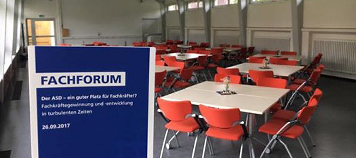 Veranstaltungsraum an der FH Münster mit Veranstaltungshinweis auf blauem Schild im Vordergrund und Tischgruppen mit orangenen Stühlen 