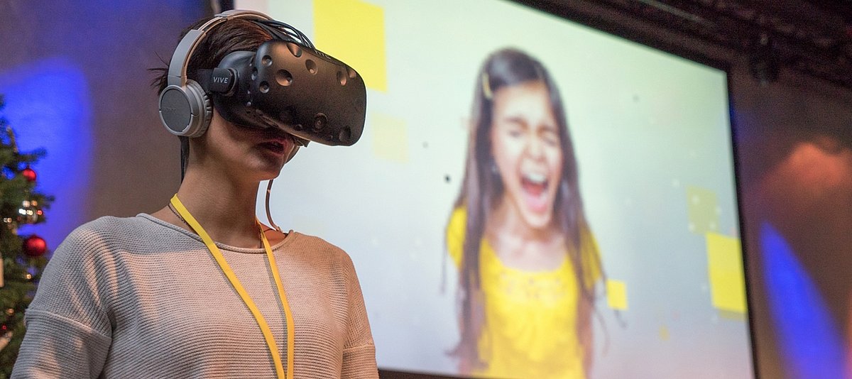Eine Frau trägt eine Virtual Reality-Brille, im Hintergrund ist ein schreiendes Mädchen auf einer Leinwand abgebildet