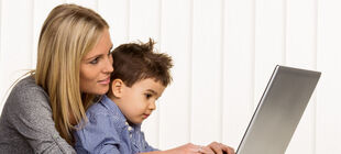 Eine Mutter hat ihren Sohn im Kindergartenalter auf dem Schoss und schaut gemeinsam mit ihm auf einen Laptop und lächelt.