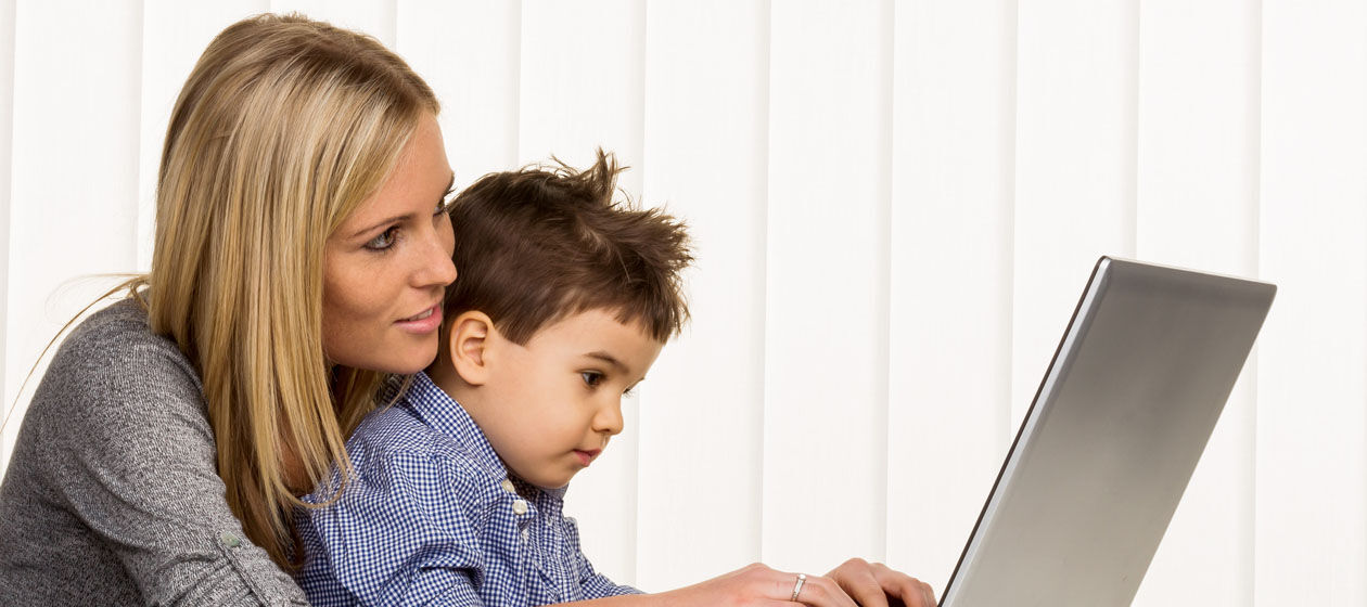 Eine Mutter hat ihren Sohn im Kindergartenalter auf dem Schoss und schaut gemeinsam mit ihm auf einen Laptop und lächelt.
