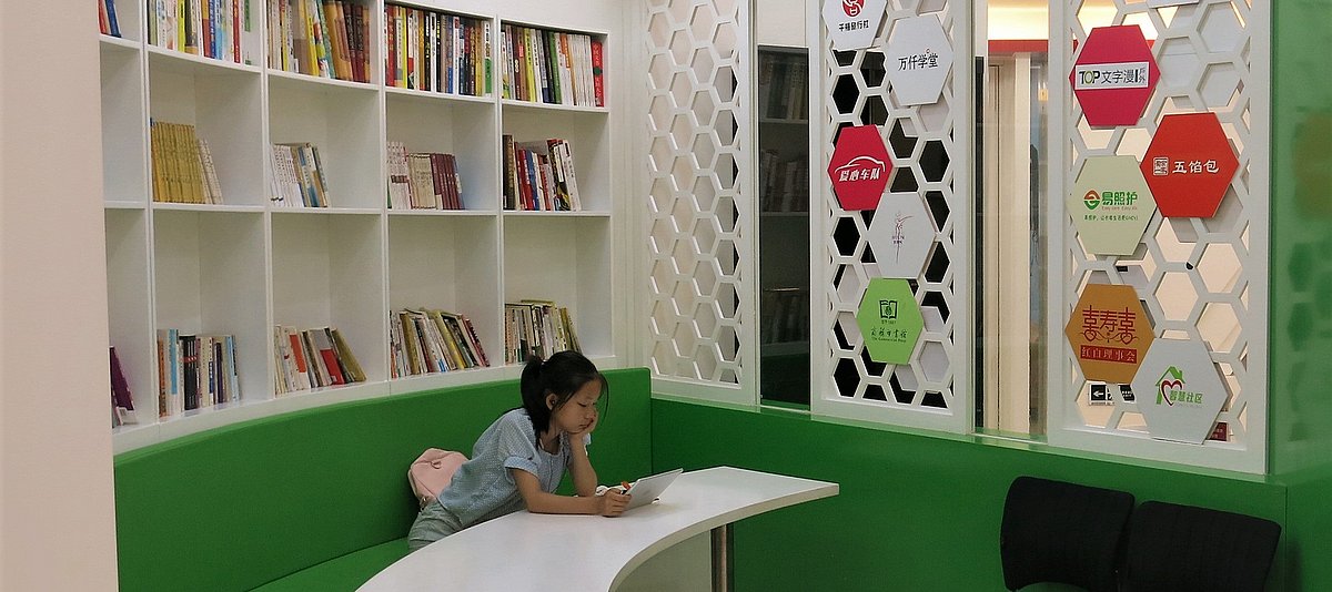 Ein Mädchen sitzt auf einer grünen Bank in der Lernecke an einem Tisch und liest auf einem Tablet. Im Halbrund hinter ihr befinden sich Regale mit Büchern, an der Wand ggü. hängen rautenförmige Poster mit chinesischen Schriftzeichen.  