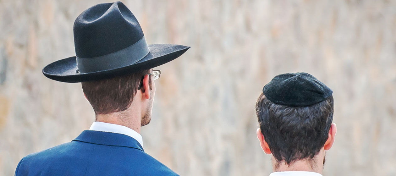 Zwei Hasedim, Männer einer jüdischen Glaubensgemeinschaft, sind von hinten abgebildet und tragen traditionelle Kopfbedeckungen.