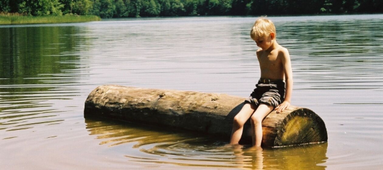 Ein Junge sitzt auf einem Baumstamm, der in einem See liegt.