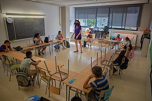 Kinder mit Mundschutz sitzen in einem Klassenzimmer und jeweils zwei leeren Plätzen zwischen ihren Sitzplätzen, Die Lehrerin steht in der Mitte der Tische.