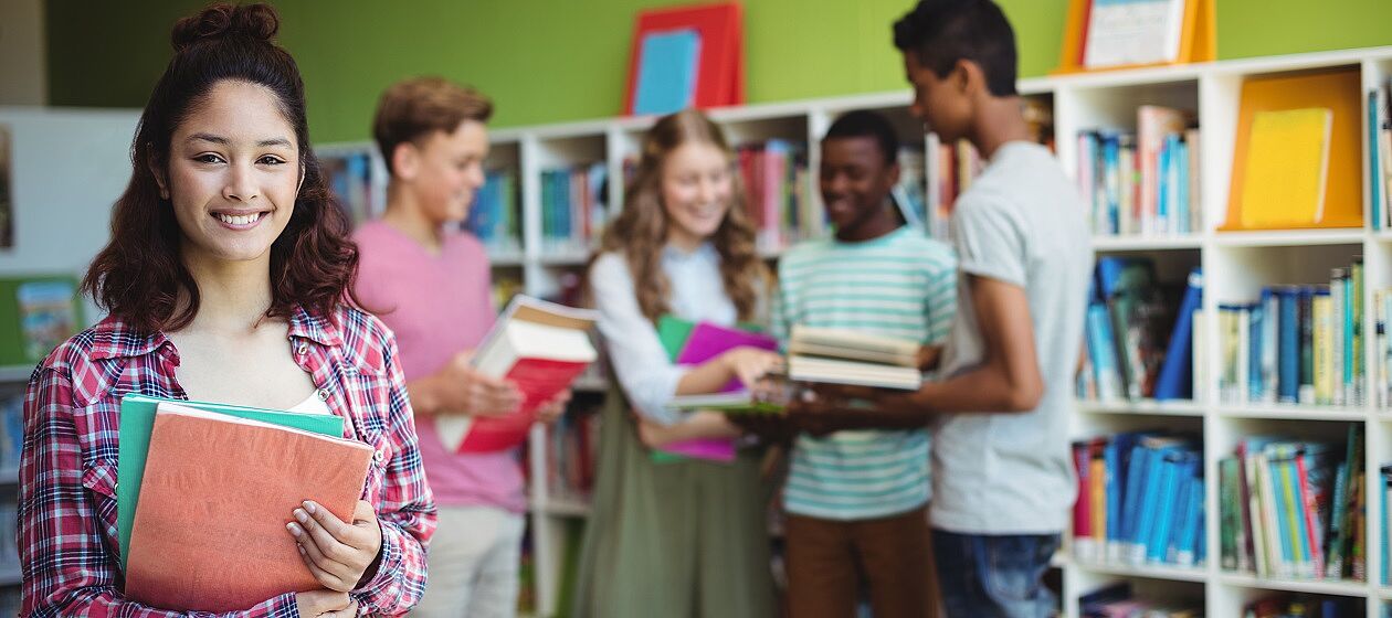 Mehrere Jugendliche mit Büchern in den Armen stehen vor einem Bücherregal, eine Person lächelt in die Kamera