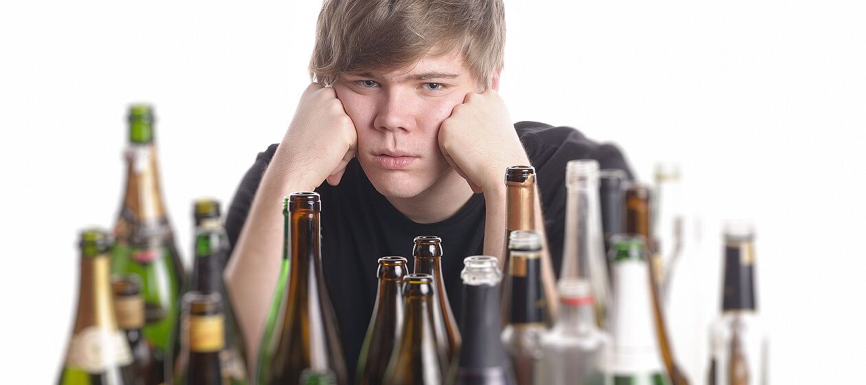 Ein Jugendlicher sitzt verzweifelt vor einer Reihe Sprirituosen-Flaschen