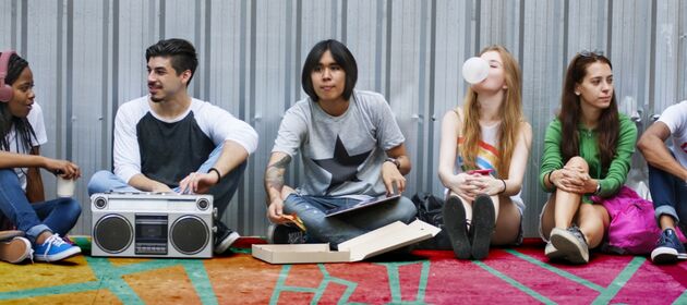 Eine Gruppe junger Erwachsener sitzt auf dem Boden und verfolgt unterschiedliche Aktivitäten wie Musik hören, Kaugummi kauen, Pizza essen,