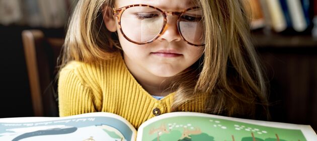 Ein Mädchen mit einer großen Brille schaut konzentriert in ein Kinderbuch