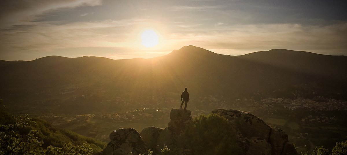 Mensch steht auf Steinhügel und schaut zur strahlenden Sonne über den Bergen