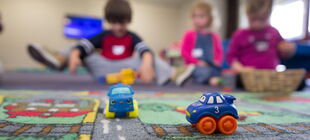 Auf einem Spielteppich sind zwei Spieulzeugautos zu sehen, im Hintergrund drei kleine Kinder die nebeneinander auf dem Boden sitzen